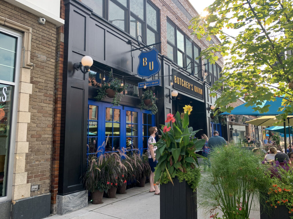 The exterior of Butcher's Union in downtown Grand Rapids on a sunny summer day, featuring outdoor patio seating, vibrant landscaping, and the restaurant's iconic blue entrance.